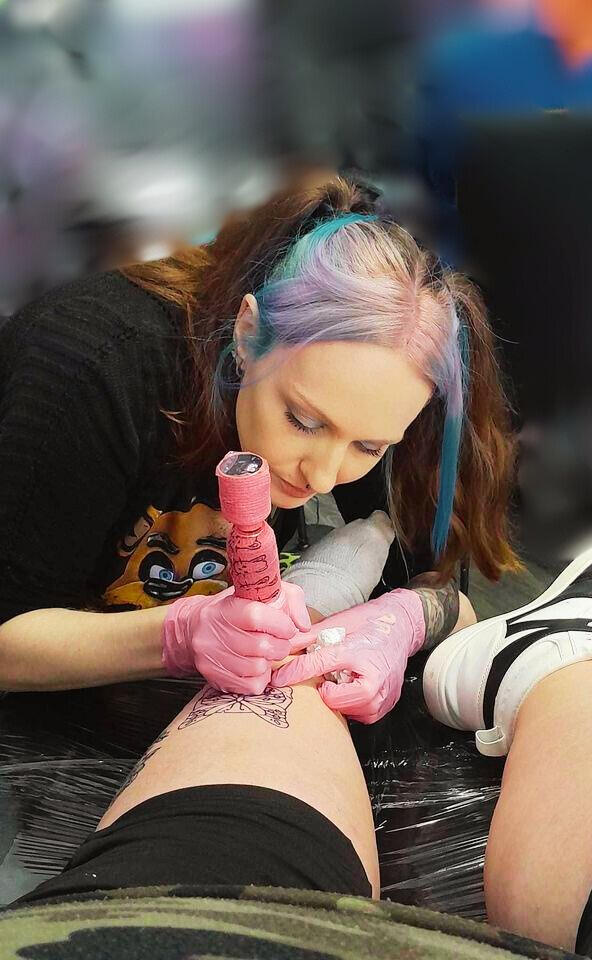 Jenn Cara (LOGO) Jenn Cara, a tattoo artist with purple and blue hair, focused on tattooing a client's leg. She is wearing pink gloves and using a tattoo machine.